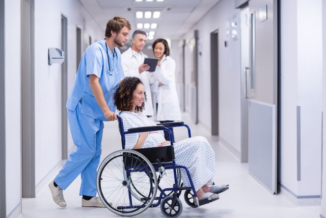 Doctor pushing a pregnant woman in a wheelchair through a hospital corridor. Medical staff in the background discussing patient care. Useful for topics related to healthcare, maternity care, hospital services, and medical assistance.