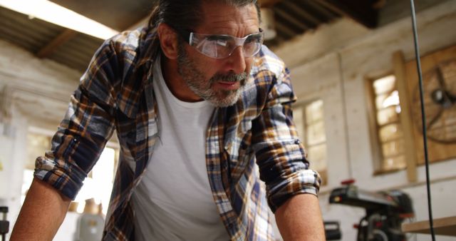 Mature man in plaid shirt and safety glasses leaning forward while working in a workshop. Perfect for visually representing skilled labor, craftsmanship, woodworking, and industrial safety themes. Suitable for use in articles, advertisements, or blog posts related to DIY projects, skilled trades, and workplace safety.