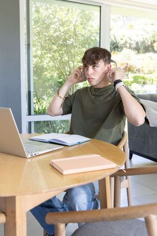 Teenager adjusting headphones while studying at home with laptop and notebook in daylight