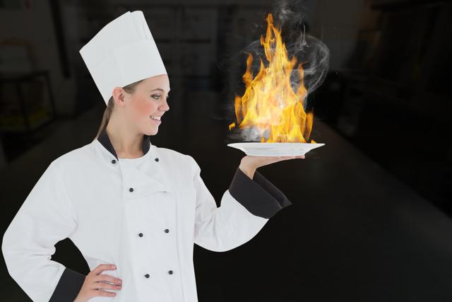 Image depicts female chef dressed in white uniform and hat, confidently holding a flaming plate. Suitable for culinary arts promotions, restaurant advertisements, cooking classes, kitchen safety materials, and gastronomy blogs. Conveys professionalism, culinary expertise, and dynamic cooking environment.