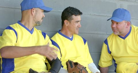 Baseball teammates sitting in dugout wearing yellow and blue uniforms holding glove