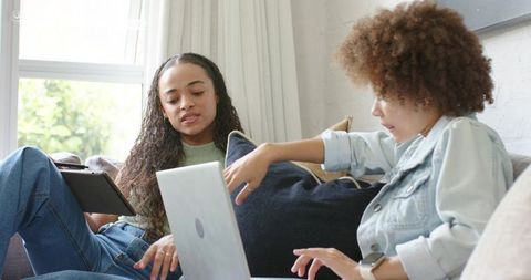 Female Friends Collaborating on Laptop and Tablet on Sunlit Living Room Sofa