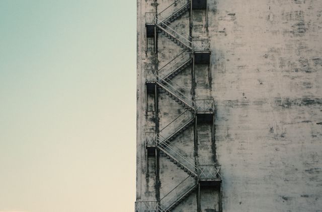 This image captures a side view of a weathered concrete building with a metal fire escape stairwell attached to it, showing the rugged texture and urban industrial setting. Ideal for articles or content about urban architecture, city safety features, emergency preparedness, or flexible space planning. Useful in a wide range of topics including safety regulations, architectural styles, or the adaptation of urban infrastructure.