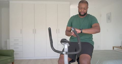 African man checking smartwatch while riding exercise bike in minimalist home bedroom