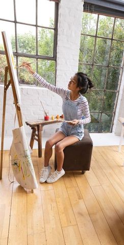 Young chinese painter working on canvas in sunlit studio wearing striped tee overalls