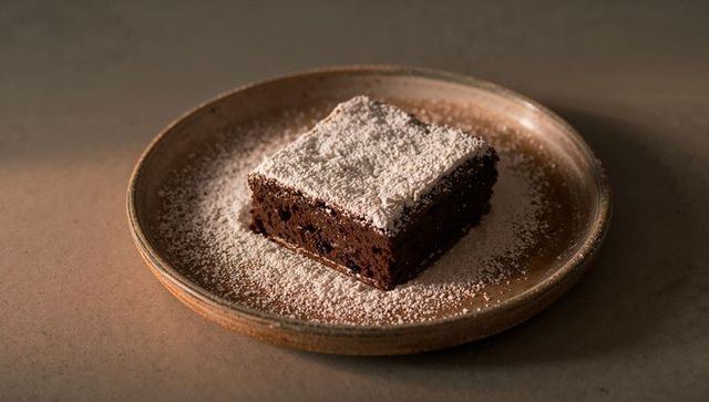 Chocolate brownie sitting on rustic ceramic plate with powdered sugar dusting