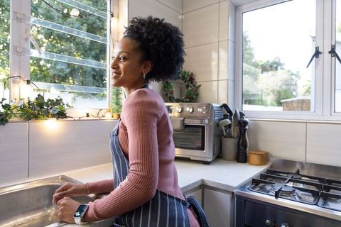 Smiling woman washing dishes by sunny kitchen window with holiday lights and cozy greenery