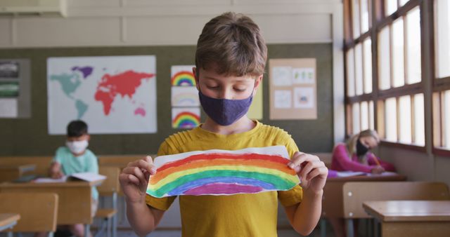 Young boy showing rainbow artwork in classroom while wearing a mask, indicates educational activities and safety protocols during COVID-19. Useful for themes on education, pandemic safety, creative activities, and social distancing in schools.