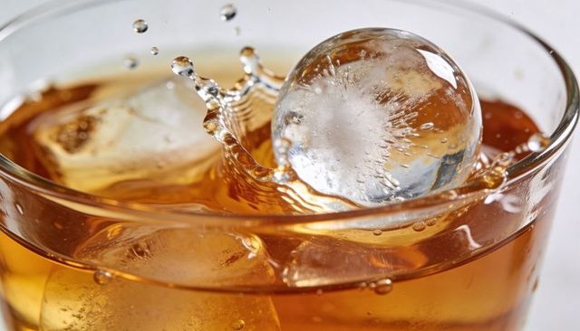 Splashing ice sphere in amber whiskey glass creating crown droplets and glossy reflections