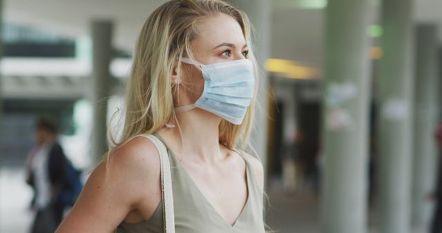 Woman with blonde hair wearing protective face mask standing outside in public area. Perfect for use in health and safety campaigns, articles about COVID-19, and illustrations emphasizing the importance of protection in public spaces.