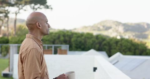 Mature bald man standing on balcony holding ceramic mug and watching peaceful hillside view