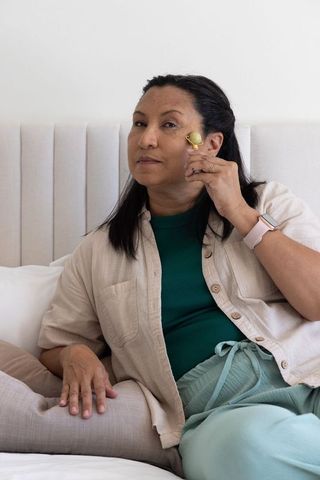 Senior african american woman massaging face with green facial roller relaxing in bed