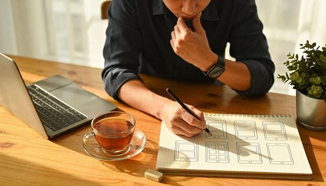 Ux designer sketching mobile app wireframes on sketchbook at desk with laptop and sunlight