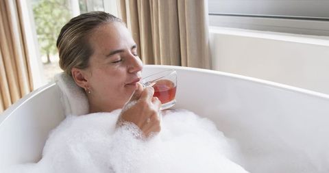 Woman relaxing in bubble bath sipping herbal tea by window in bright modern bathroom