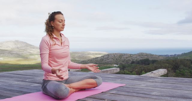 Woman is sitting in lotus position on yoga mat, meditating. Background includes mountains and ocean under a clear sky. Useful for promoting wellness, mental health, yoga retreats, relaxation techniques, well-being.