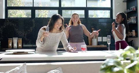 Multicultural women laughing and chatting around bright modern kitchen island with drinks