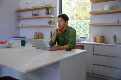 Asian man working on laptop at modern kitchen island holding pen thoughtful home office