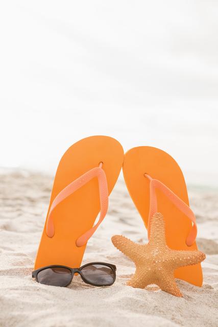 Orange flip flops standing upright in sand next to sunglasses and starfish on a beach. Ideal for summer vacation promotions, travel brochures, beachwear advertisements, and holiday-themed social media posts.