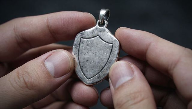 Hands holding worn shield pendant showing patina, engraved crest and bail loop closeup