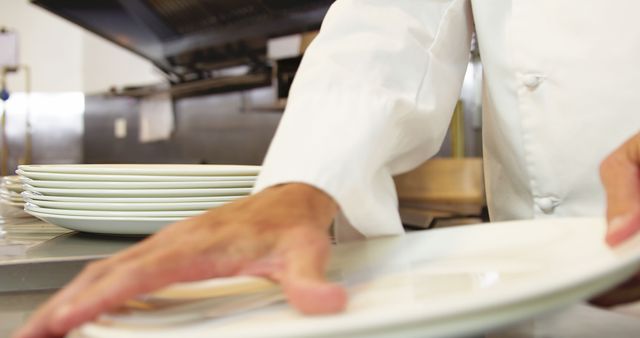 Chef wearing white uniform setting stack of clean plates in a professional kitchen. Useful for blogs or articles about culinary arts, restaurant preparation, professional cooking environments, or food industry operations.