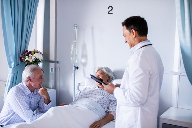 Senior patient lying in hospital bed, interacting with doctor holding a clipboard. Another senior person, possibly a family member, sitting beside the bed. Ideal for use in healthcare, medical consultation, elderly care, and hospital-related content.