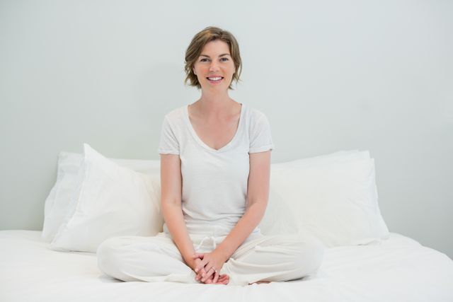 This image shows a young woman sitting cross-legged on a bed in a bright bedroom. She is smiling and appears relaxed, dressed in casual white clothes. The setting is serene and comfortable, making it ideal for use in lifestyle blogs, wellness websites, home decor promotions, and advertisements focusing on relaxation and comfort.