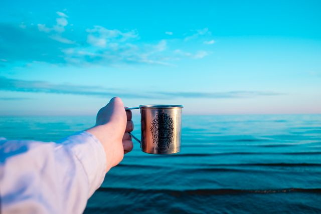 Hand holding a metallic cup towards calm blue sea during sunset. Ideal for travel blogs, adventure websites, camping gear advertisements, and inspirational posters.