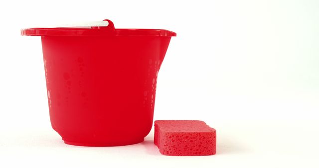 A red plastic bucket with water droplets is paired with a pink cleaning sponge, with copy space. These items are commonly used for household chores and cleaning tasks.