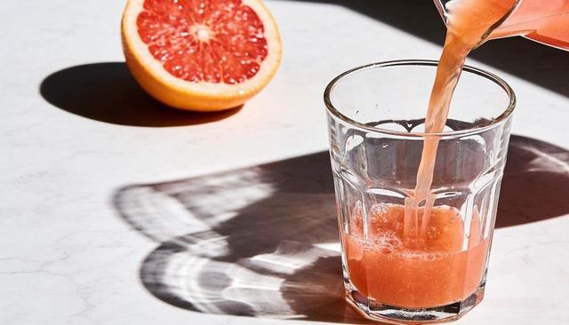 Pouring pink grapefruit juice into faceted glass on white marble with sunlit shadow