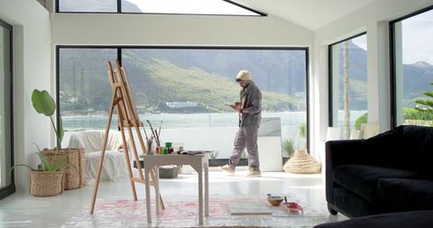 Indian artist standing by easel in contemporary waterfront studio with mountain view