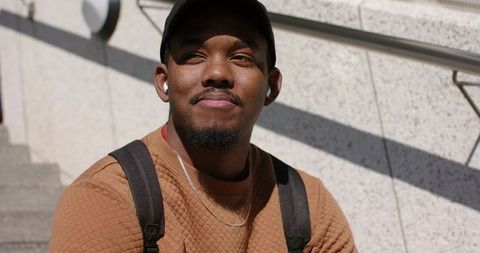 African american man sitting on urban steps listening to earbuds wearing backpack and brown sweater