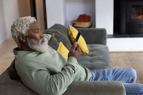 Senior african american man reclining on gray sofa using smartphone by fireplace, relaxed home tech