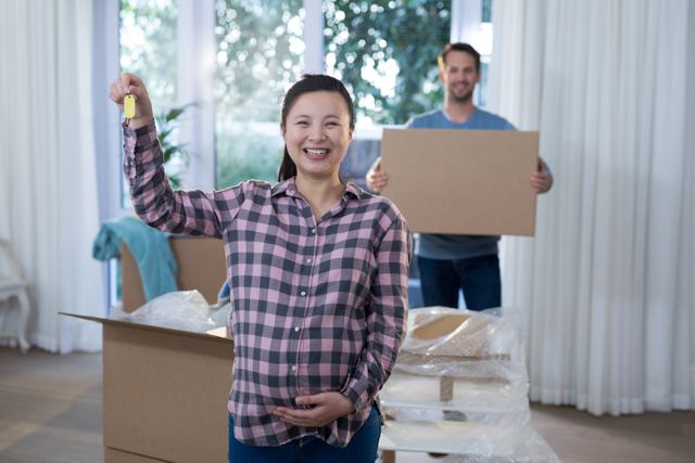 Pregnant woman smiling and holding a key while her partner carries a box in the background. Ideal for use in real estate promotions, family lifestyle blogs, and advertisements about moving or homeownership.