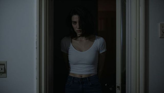 Woman standing in dim hallway wearing white crop top and jeans casting dramatic shadow