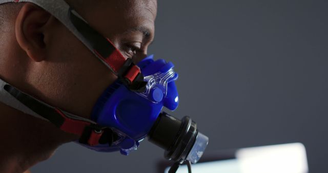 A close-up of a man wearing a gas mask with blue filters, showing dedication to health safety measures indoors. Suitable for use in content related to pandemics, environmental health, industrial safety, or respiratory protection topics.