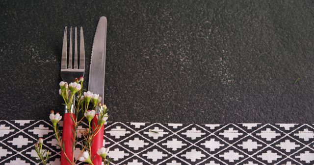 Fork and knife with red handles placed on patterned tablecloth against a dark background. Adorned with small white flowers adding a decorative touch. Ideal for use in content about dining, table settings, party setups, and food presentation ideas.