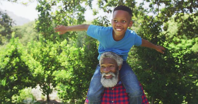 Happy african american grandfather carrying smiling grandson on his shoulders outdoors in nature. Family, fun, nature and togetherness.