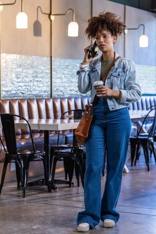 African woman walking in cafe talking on phone holding coffee to-go wearing denim jacket