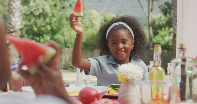 African American girl sitting at a table outdoors with family, joyfully holding up a slice of watermelon. The scene suggests a summer picnic, family vacation or weekend relaxation. Great for advertisements focusing on family gatherings, outdoor leisure activities, summer promotions, healthy eating, and childhood happiness.