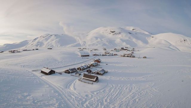 Windswept alpine hamlet with wooden cabins and smoking chimneys on expansive snowfield