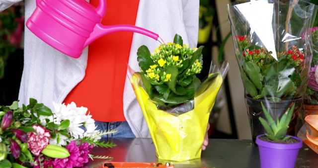 Person watering a vibrant yellow flower pot with pink watering can in a floral shop. Colorful floral arrangements and gardening tools visible in the background. Ideal for advertisements related to gardening, home decor, floral shops, and botany-themed blogs.