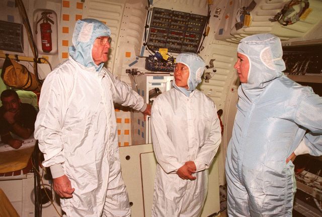 Three astronauts in cleanroom suits engaged in discussion about mission payloads inside a spacecraft. The image features significant preparation activities related to the STS-95 mission aboard the Space Shuttle Discovery, launched in 1998. Useful for articles or educational material about space mission preparations, historical space missions, NASA, and astronaut training procedures.