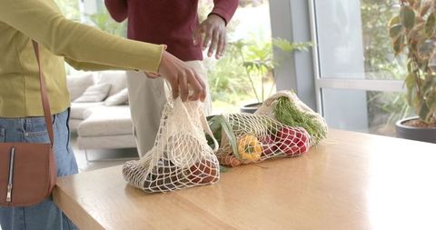 Diverse couple unpacking reusable mesh bags with fresh vegetables on sunlit wooden table