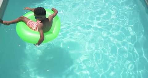 African american boy floating on neon tube in turquoise pool wearing sunglasses and pink trunks