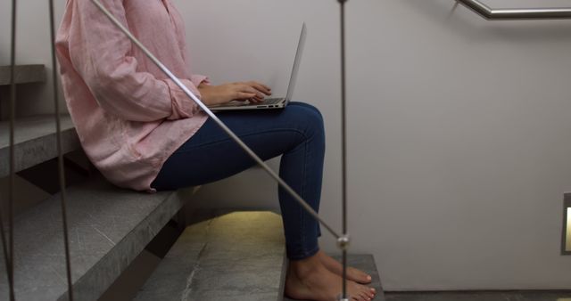 Person wearing a pink shirt and jeans sitting on stairs while working on a laptop. Ideal for representing remote work, freelancing, and work-from-home scenarios. Useful for articles on productivity, unconventional workspaces, or modern working environments.