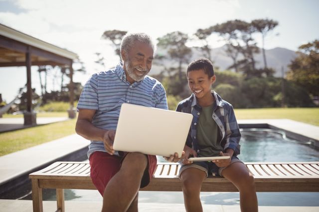 Smiling grandfather showing laptop to grandson near poolside on a sunny day