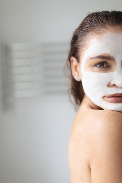 This image of a woman applying a facial mask in her bathroom can be used in content related to skincare routines, beauty and wellness tips, home spa treatments, self-care practices, and cosmetic product advertisements. It highlights relaxation and personal care.