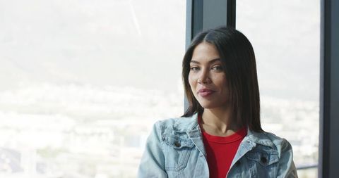 Confident Indian woman standing by highrise window with city skyline, wearing denim jacket