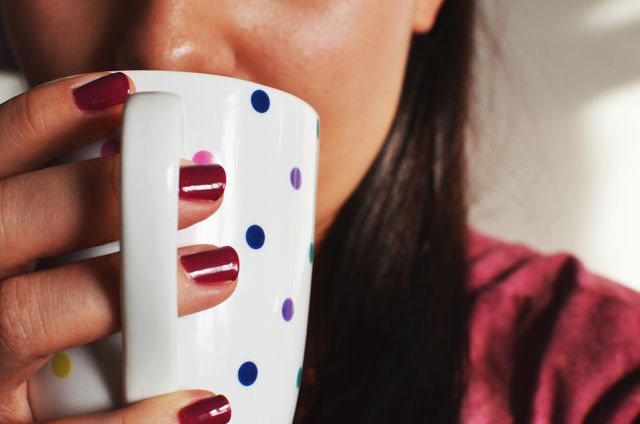 This image shows a close-up of a woman holding a white mug with colorful polka dots. Her nails are painted a vibrant color, and she is in casual clothing. This photo can be used in contexts related to lifestyle, relaxation, coffee breaks, personal care, or editorial content revolving around women's daily life.