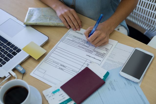 Female executive filling out a form at her office desk. The desk is cluttered with a laptop, smartphone, coffee cup, passport, travel documents, and a pen. Ideal for use in business, office, and professional work-related themes, as well as travel and administrative tasks.
